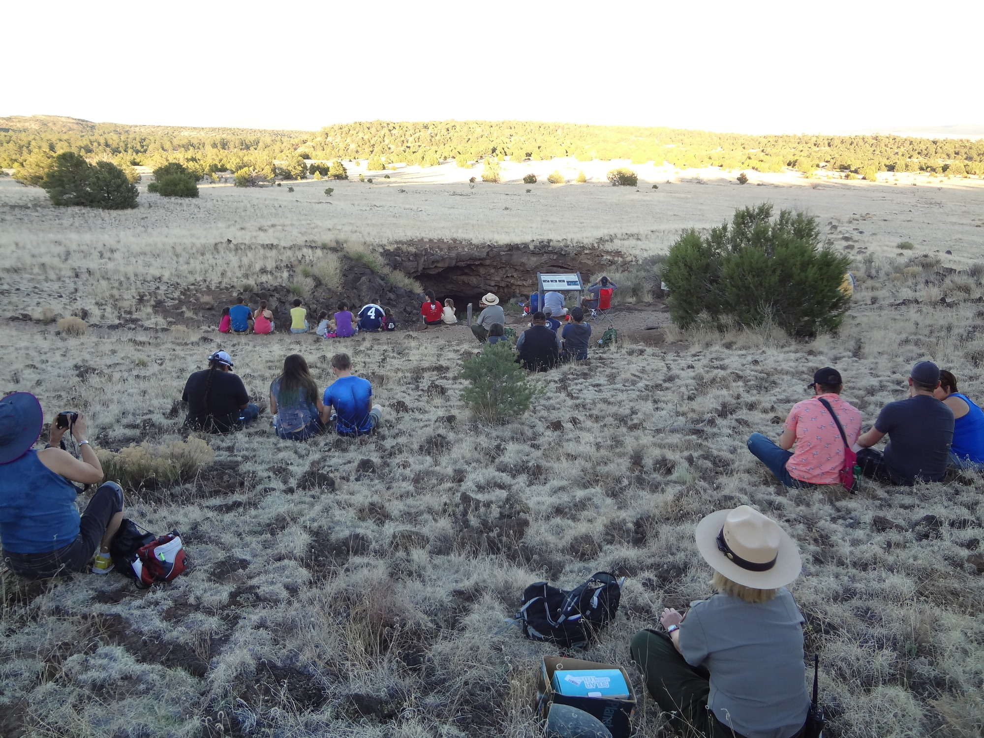 A park ranger and groups of two and three hikers sit in a grassy area.  All of them are looking at a cave entrance in front of them.