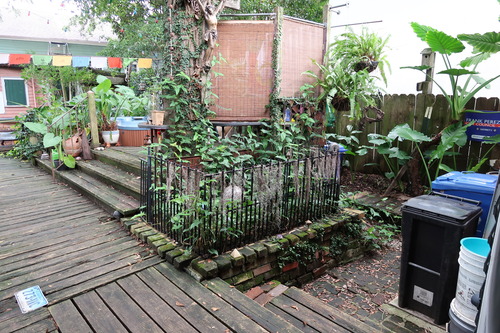 A back yard with a wooden deck with multiple levels; in the center of the photo is a garden surrounded by bricks and a cast iron fence.
