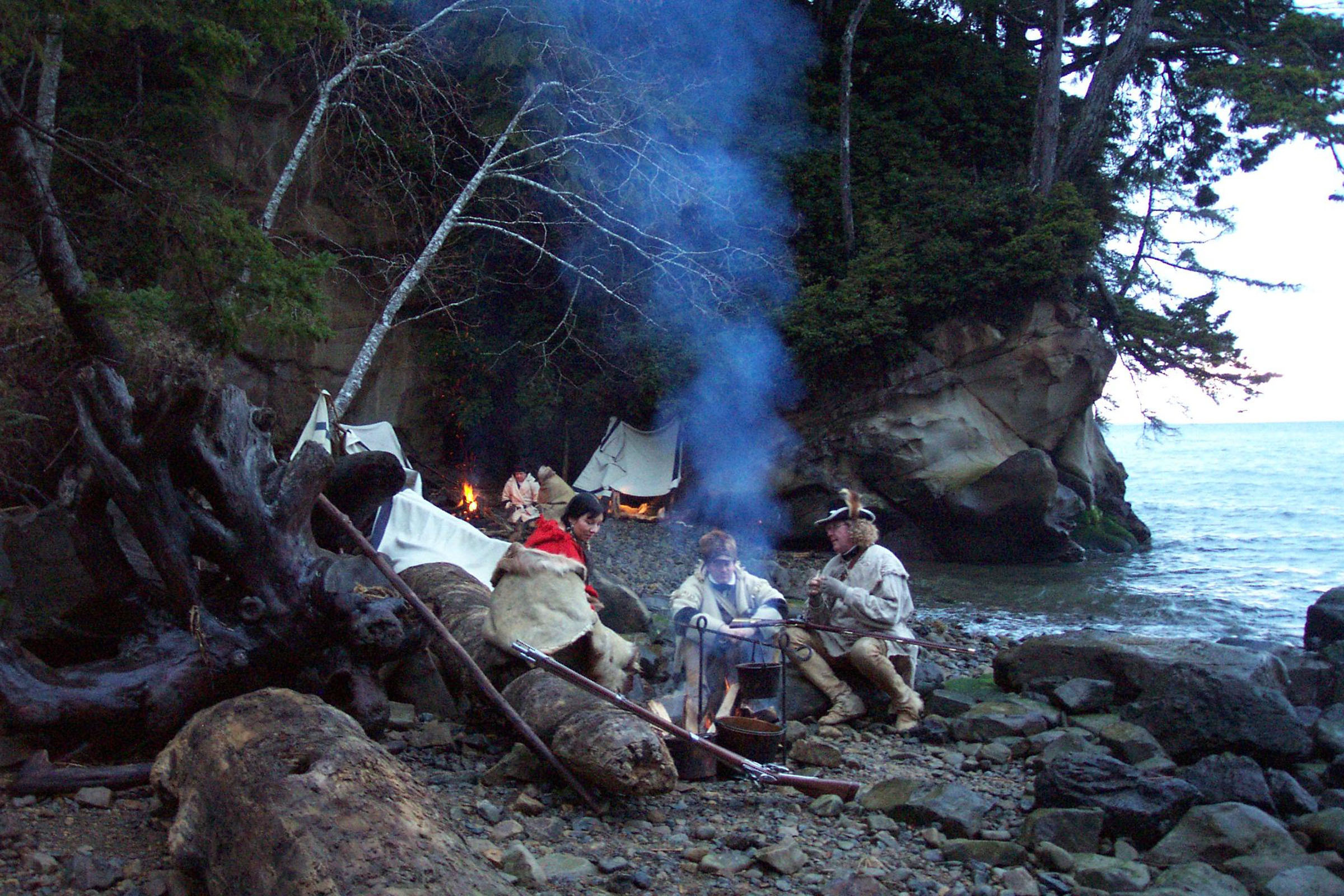 Reenactors in 1800s American attire sit around a campfire on the pacific northwest coast.