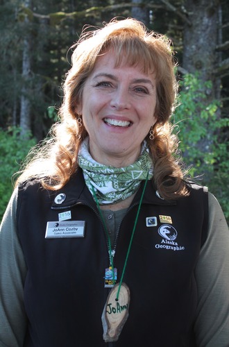 Woman smiling in a friendly manner. She wears a black vest that says "Alaska Geographic" as well as several pins.
