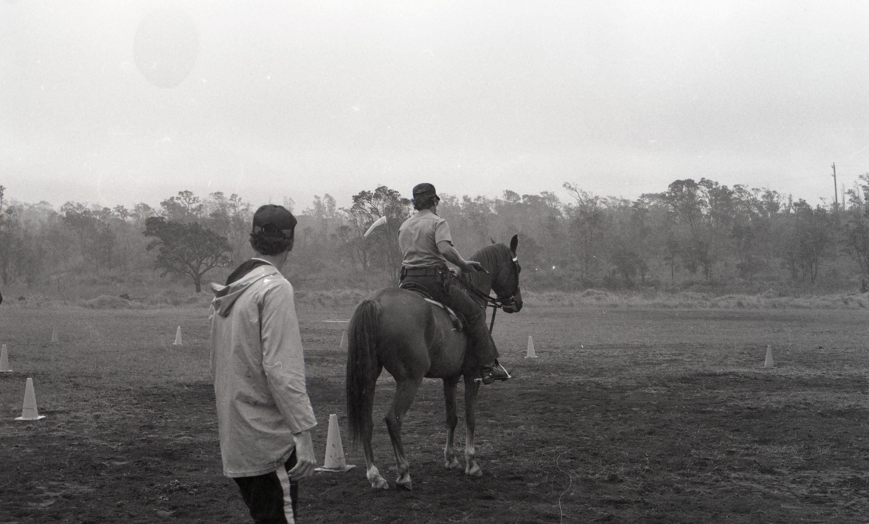 A black and white image of two men training a horse at Kilauea Military Camp. Toward the left side of the image is a man with his back towards the camera watching a man in the center of the image riding a horse. The man on horseback is also facing away from the camera. There is a cone next to the horse on its left side. There are six cones scattered across the field in the background.