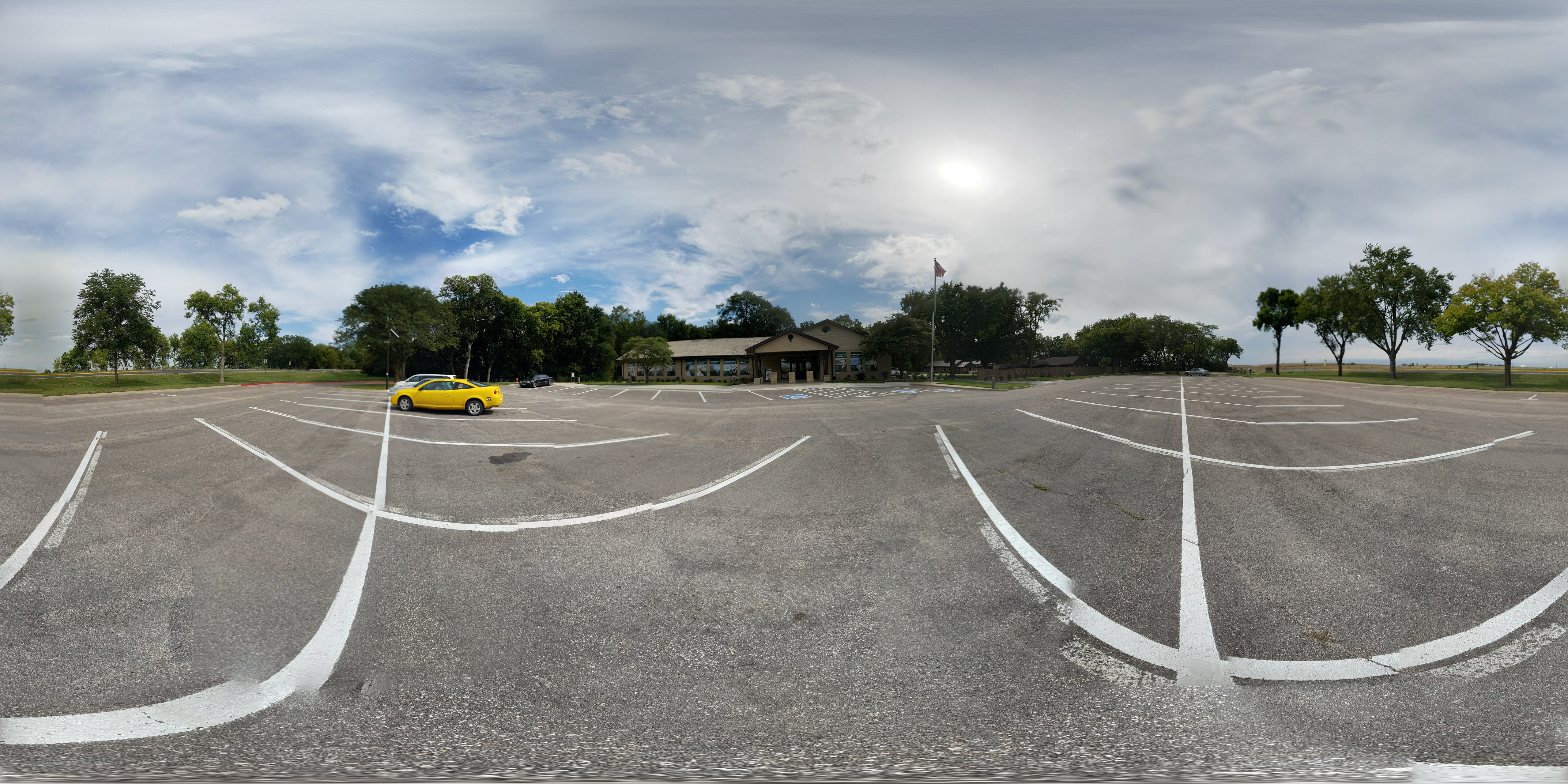 A 360-degree view of the Education Center parking lot. There is a light brown brick building covered in green banners. The parking lot has a few cars.