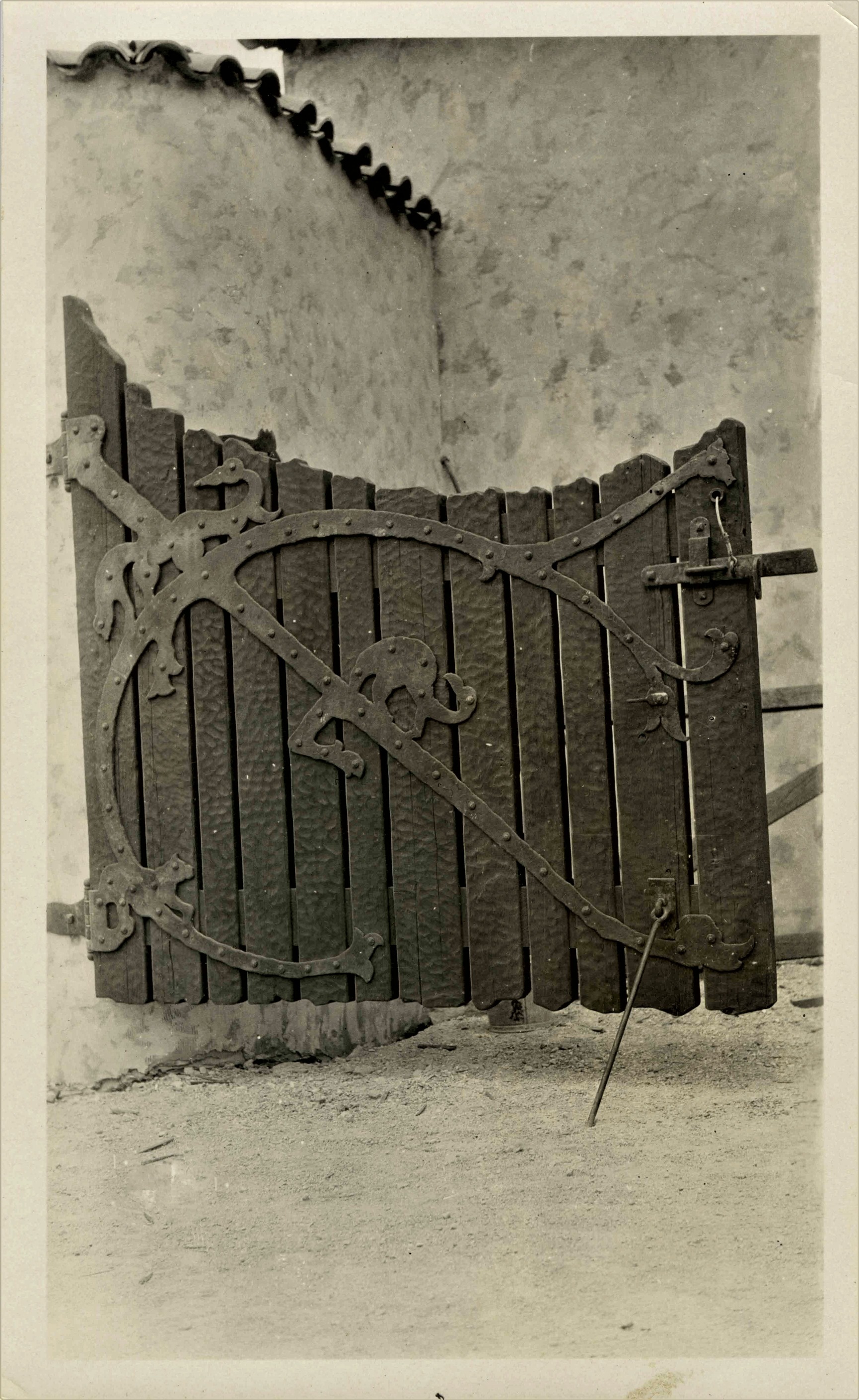 This is an historic black and white photograph from the Scotty's Castle Historic Photograph Collection, Death Valley National Park detail of one half of gate at Scotty’s Castle Stables. Gate hinged to rounded, mottled, stucco wall with flat wall behind. Gate is made of heavy, textured, vertical boards held with massive, handwrought, metal strap hinge. Hinge spans entire width of gate with stylized, desert animals. Heavy, handwrought latching system and pin at bottom to hold hate open.
