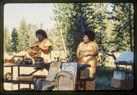 Two women stand in front of a display of Atsugewi crafts in front of a meadow. 