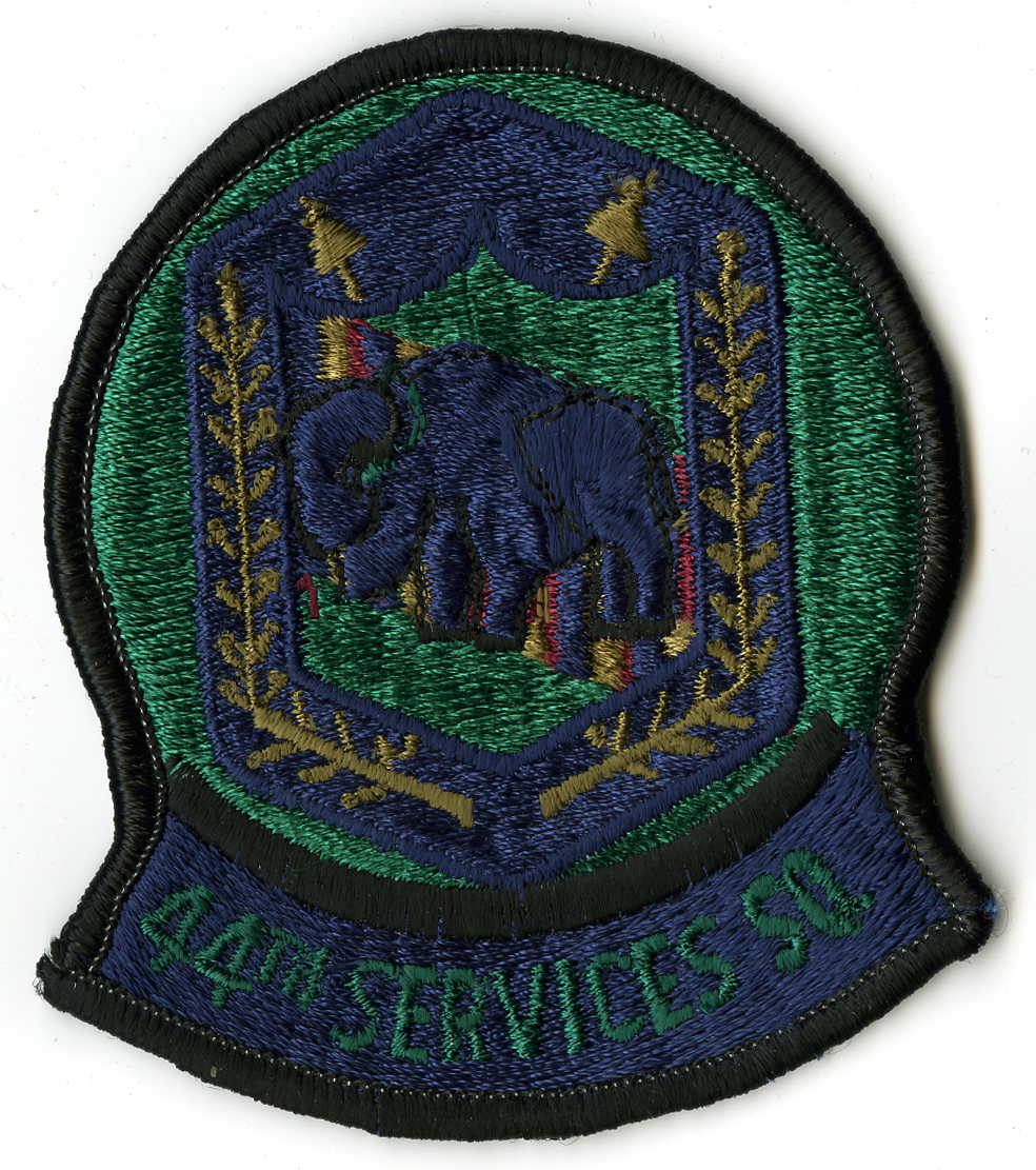 Green circular with black border and dark blue tab at bottom with 44TH SERVICES SQ" in green. In circle is blue shield with smaller green shield in center with blue bison and red  olive  and blue stripes behind. Olive branches on either side of center shield  wrapping around bottom. Top has two (2) olive arrows  one pointing away from each side of top."





