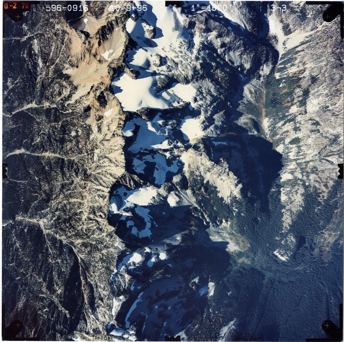Color aerial photograph of glacier and snowfields on mountain peak with forested valleys below.