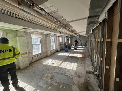 A long room with white painted brick and open windows to the left. A person stands at the far end near a ladder. Close to the camera is another person wearing a bright yellow jacket that reads 'NPS'. The floor is exposed concrete and to the right are exposed wall supports.