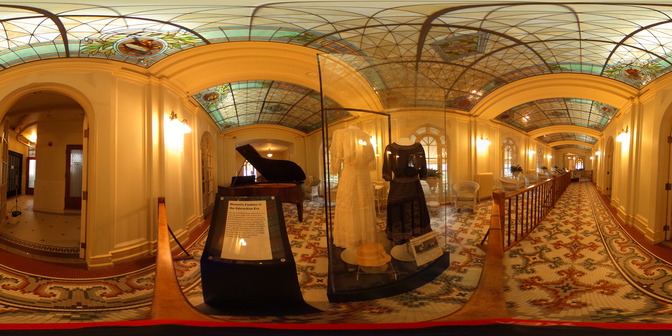 Ornate stained glass and colorful tile floors adorn this assembly room. A baby grand piano sits in the distance next to a display case featuring historical garments.  