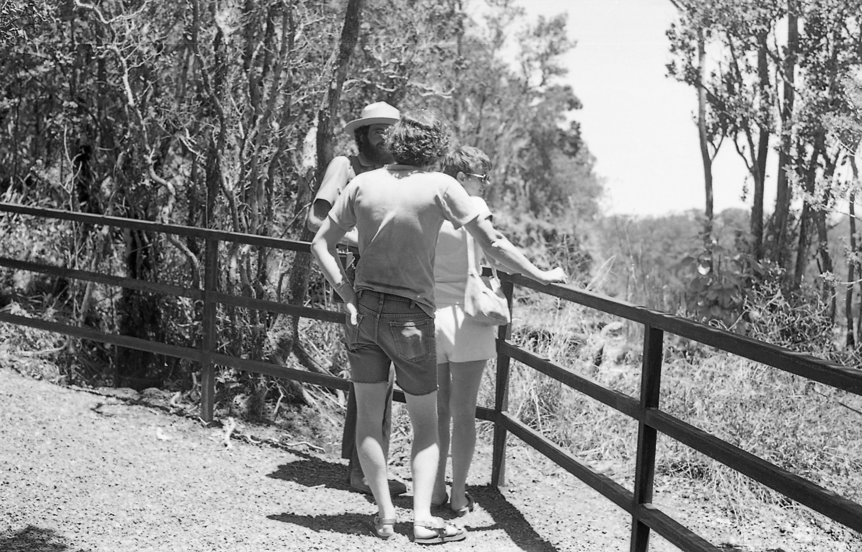 A black and white image of a male park ranger talking to visitors at a lookout along a trail. The park ranger and visitors are standing next to a wooden railing in the center of the image. The park ranger is wearing a ranger hat, a long sleeve collared shirt, pants, and boots. To the right of him is a woman looking down towards the left side of the image, she is wearing sunglasses, a short sleeve shirt, and shorts. Lastly, behind the woman is a man with curly hair. He is wearing a t-shirt, cut-off jean shorts, and sandals. His right hand is holding onto the top of the wooden railing. His left hand is in his back left pocket of his shorts. In the background behind the railing is a wooded landscape.