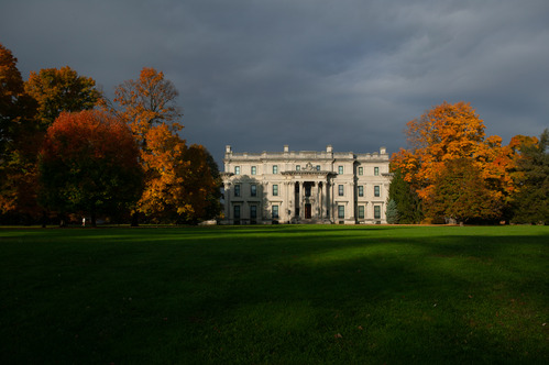 A grand three story stone house set on a vast lawn with large trees.