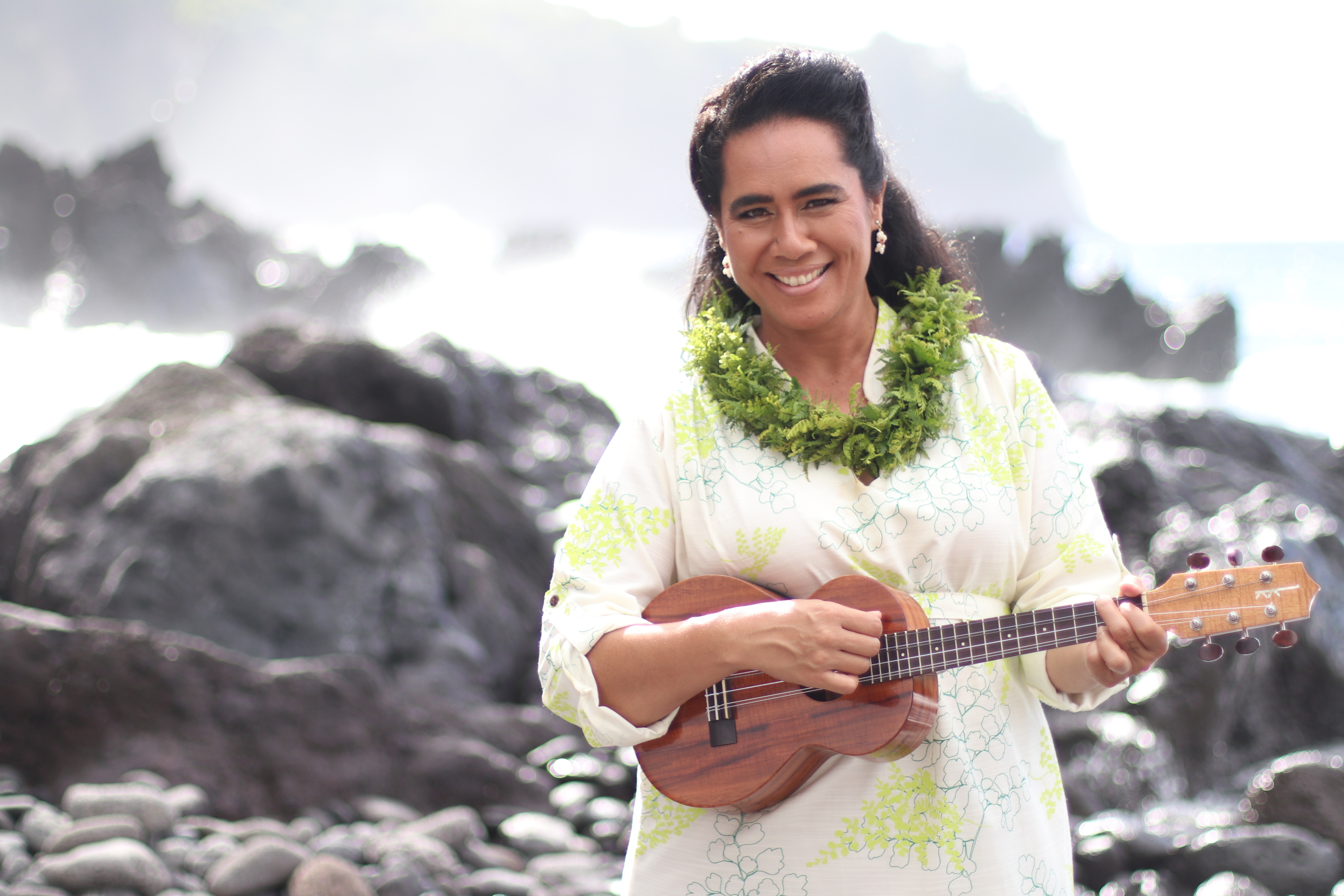 A woman smiles holding a ʻukulele near the coastline 
