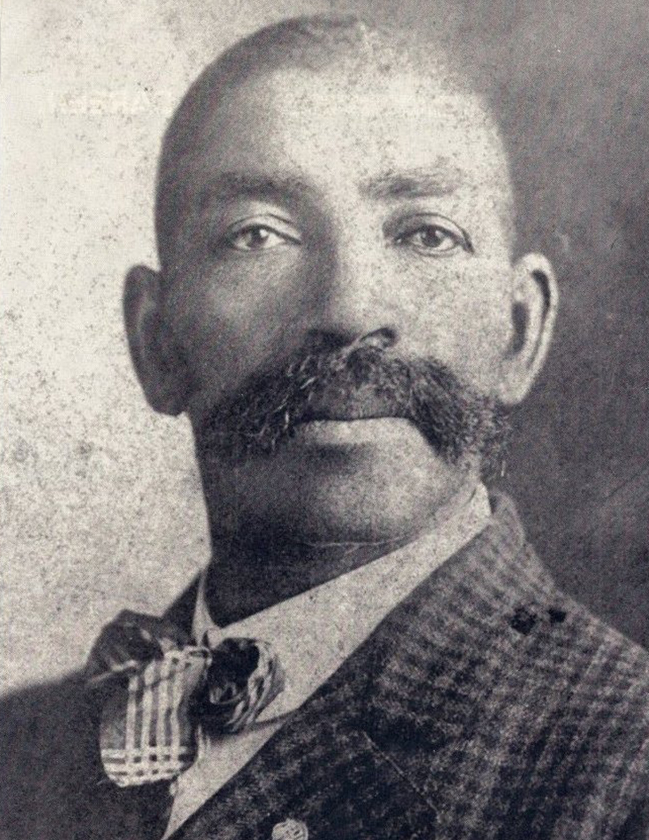 Blakc and white image of a black man with  very short hair. He has a thick mustash extending beyond his lips. He is wearing a plaid coat, light colored shirt, and a loose bowtie.
