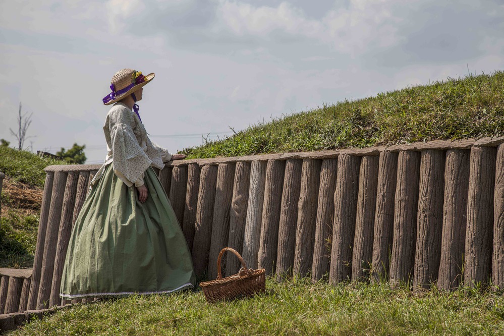 A woman in 19th-century period clothing looks over an earthwork fortification.