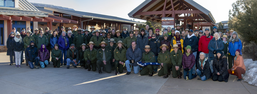 Outside, on a visitor center plaza, a group of around 75 people, wearing winter jackets, and some wearing National Park Service uniforms, are posing in several rows for a group photo at a memorial ceremony.