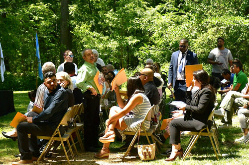50+ people fill outdoor chairs intended for participants and audience. Some are talking, shading their faces, looking around, and looking to the stage.