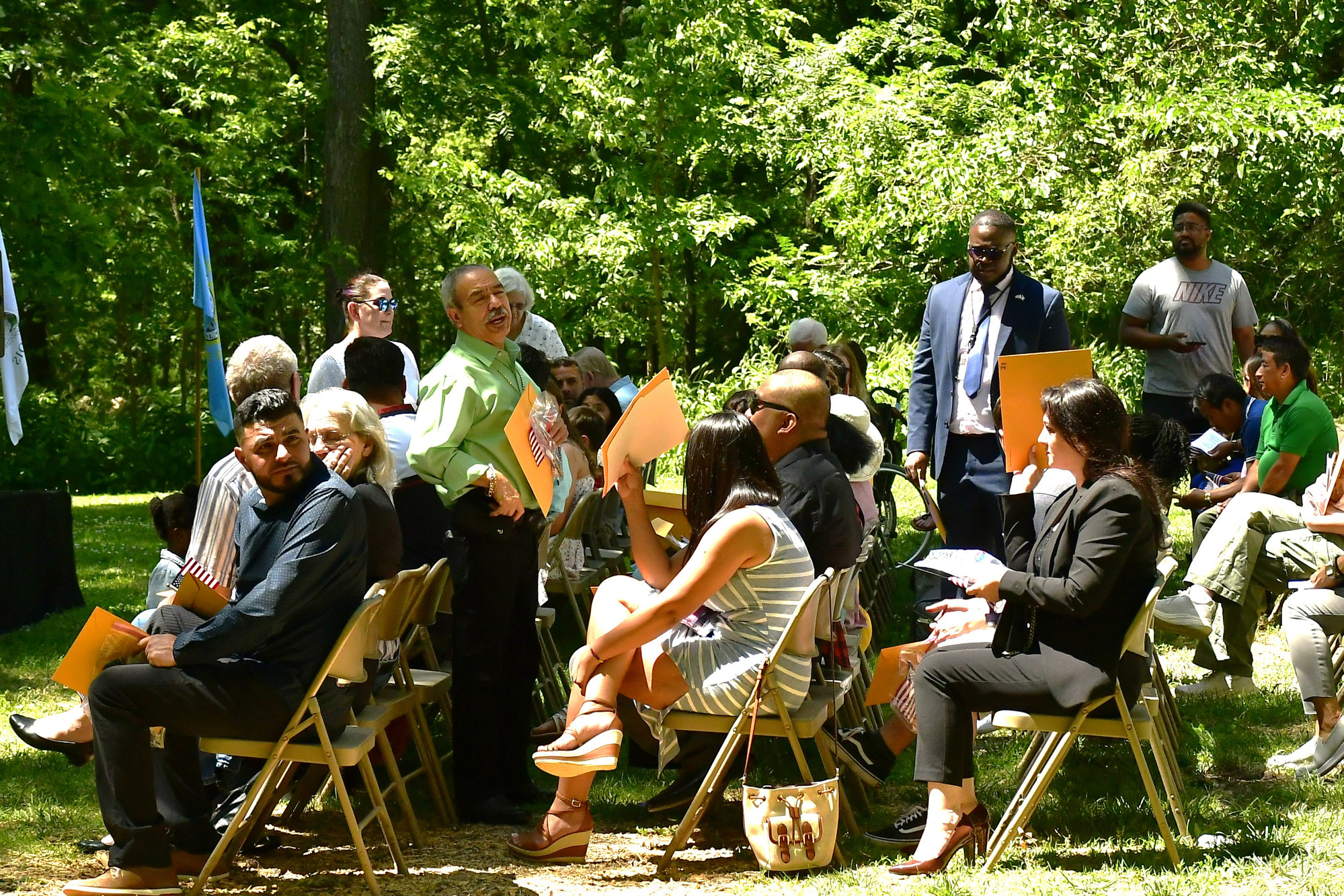 50+ people fill outdoor chairs intended for participants and audience. Some are talking, shading their faces, looking around, and looking to the stage.