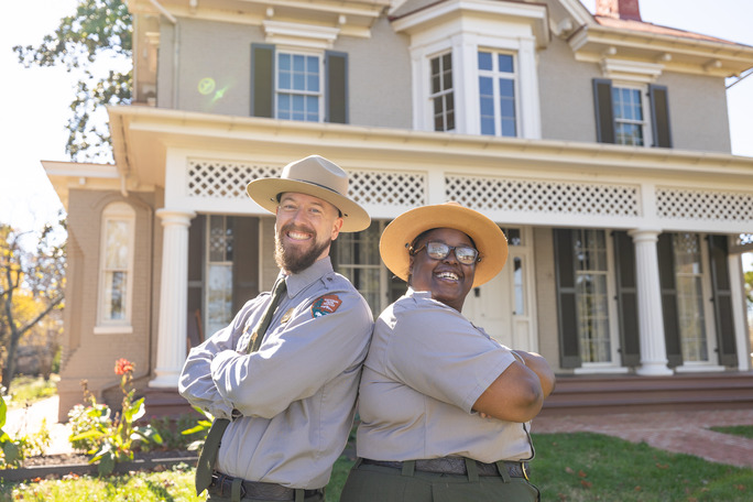 Two park rangers in uniform, a white man and a black woman, smile in front of a two story historic building.