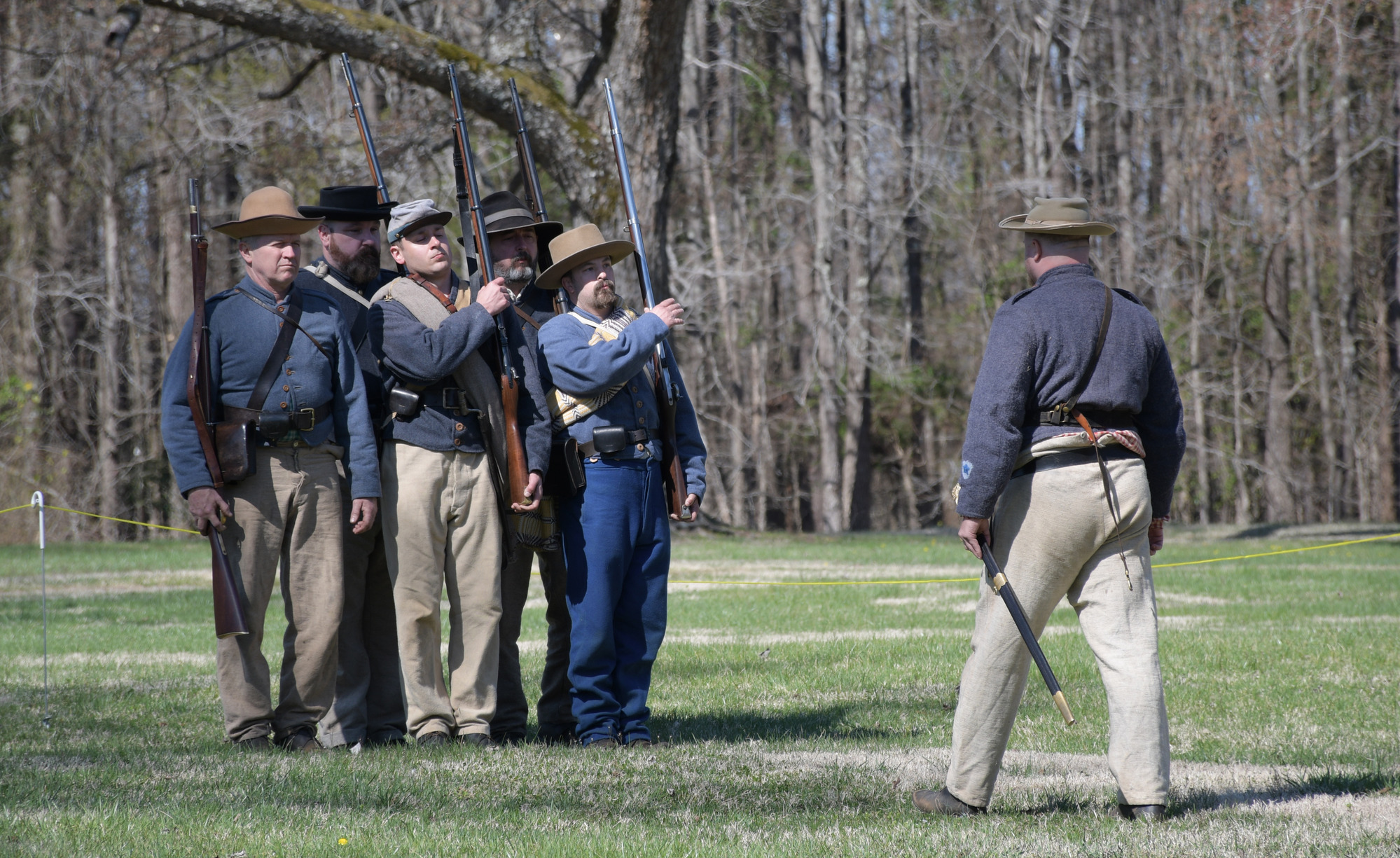 Five men wearing Confederate uniforms stand and tight formation while a fifth man stands in front of them delivering orders.