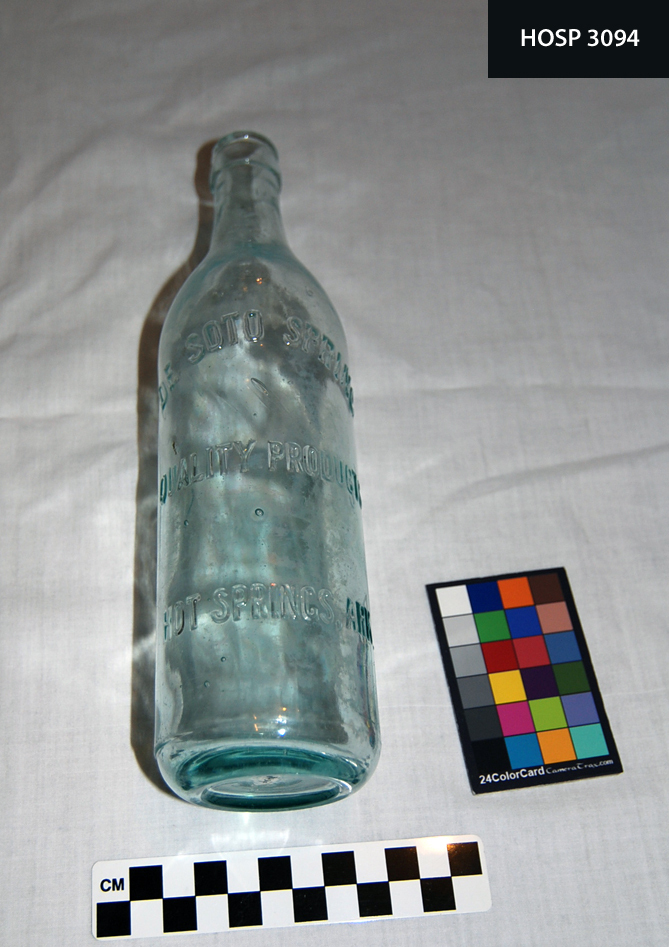 Green-tinged glass bottle for spring water. Cylindrical bottle with sloped shoulders, cylindrical neck; faint molding lines divide bottle longitudinally in half; glass band below lip; Embossed on side: DE SOTO SPRING/QUALITY PRODUCTS/HOT SPRINGS, ARK. Lip chipped