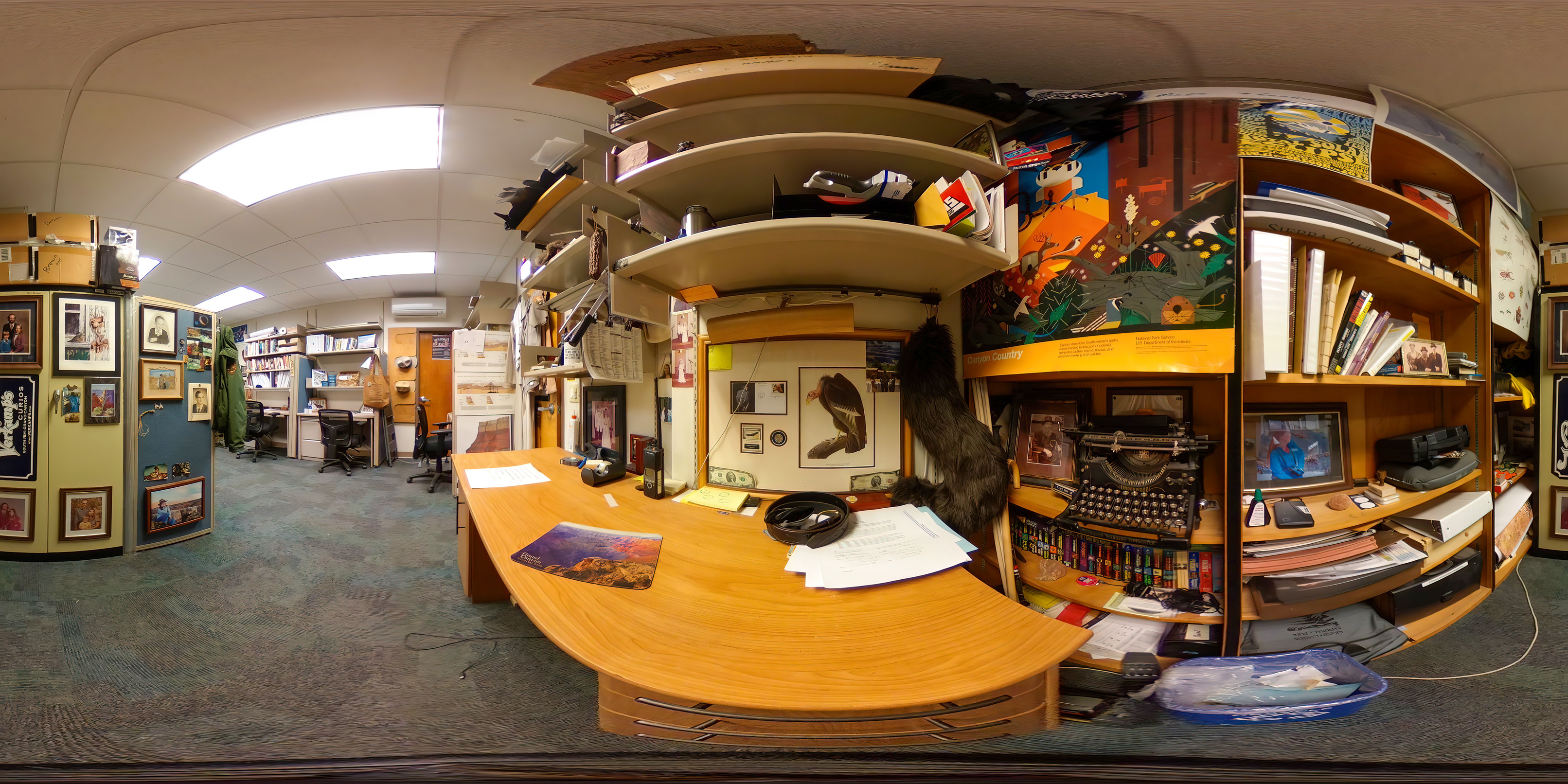 360 photo of an interpretive ranger's desk and workspace. Above the desk are book shelves. A second set of bookshelves fills the wall on the immediate right of the desk. There are books, an antique typewriter, rocks, figurines, and a variety of other nick-nacks filling the shelves. Directly behind the desk is a partition that displays about a dozen photos of the ranger and his family, from different times during his life. 