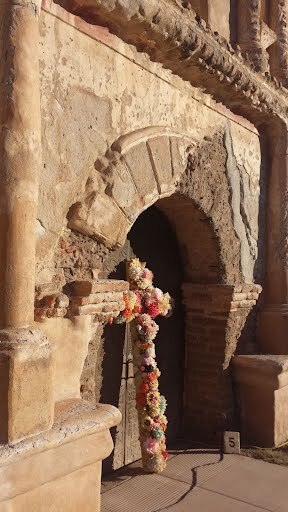 A tall cross decorated with flowers lays against an adobe mission