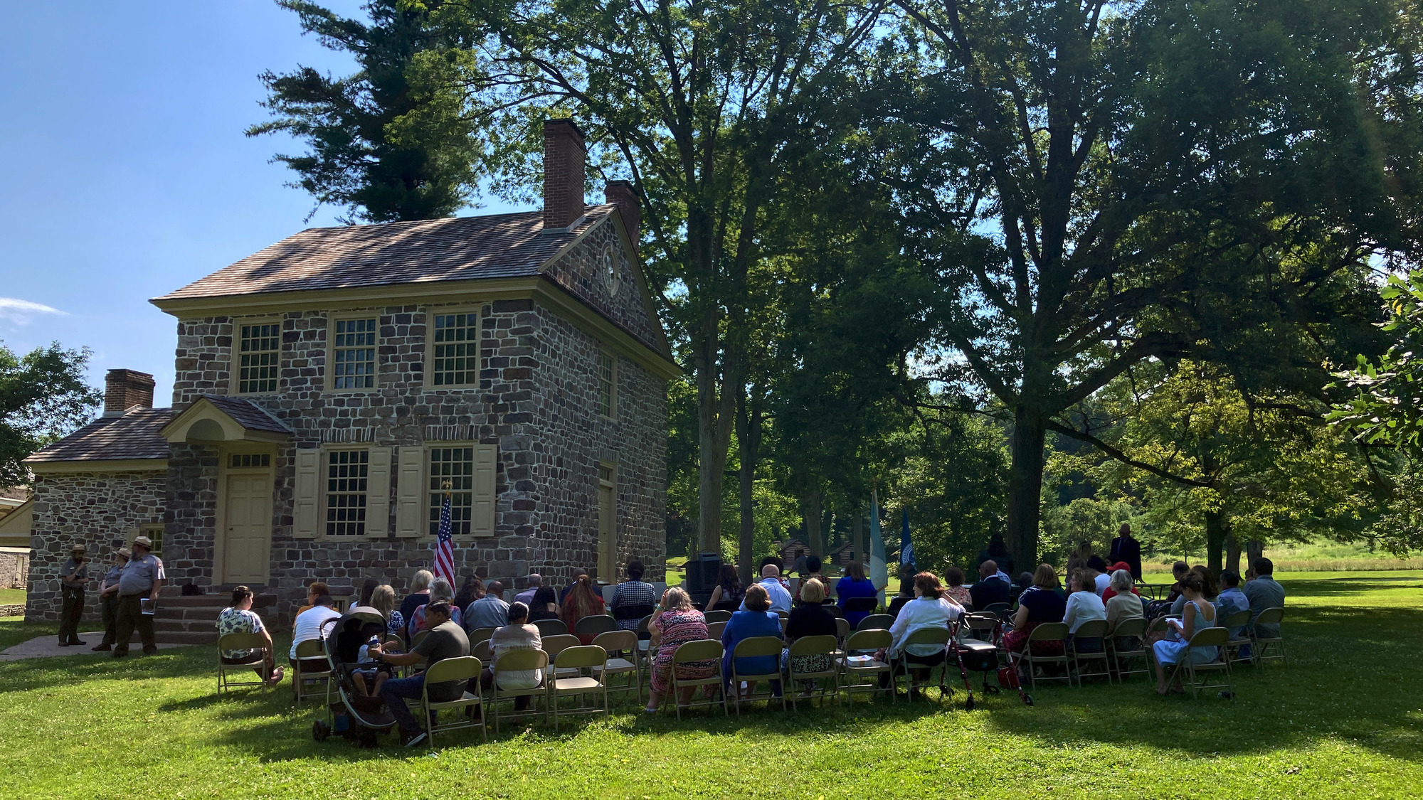 people sit in metal folding chairs in front of a small two story stone farmhouse