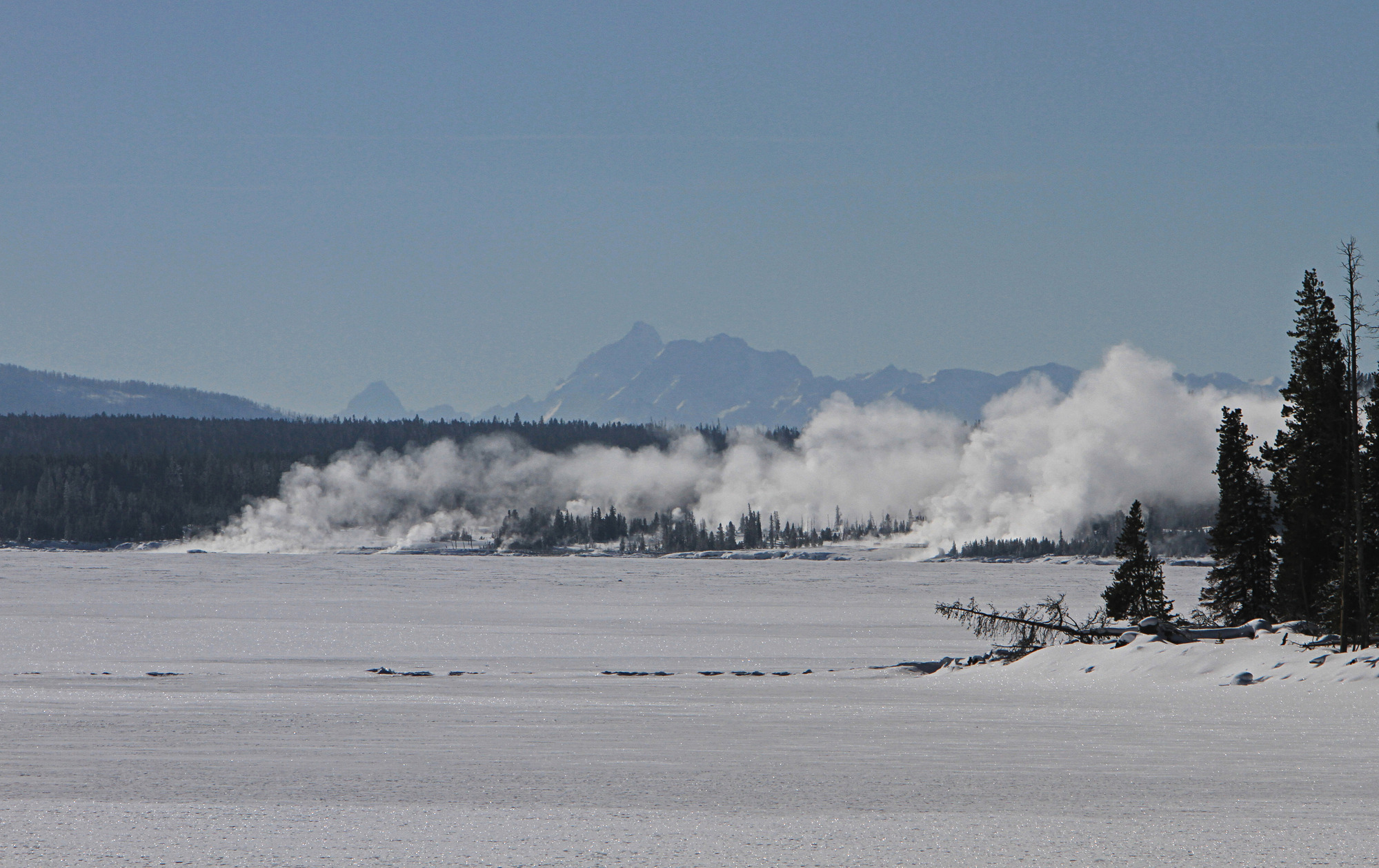 Looking across a large frozen lake at steam rising and high mountains in the background.