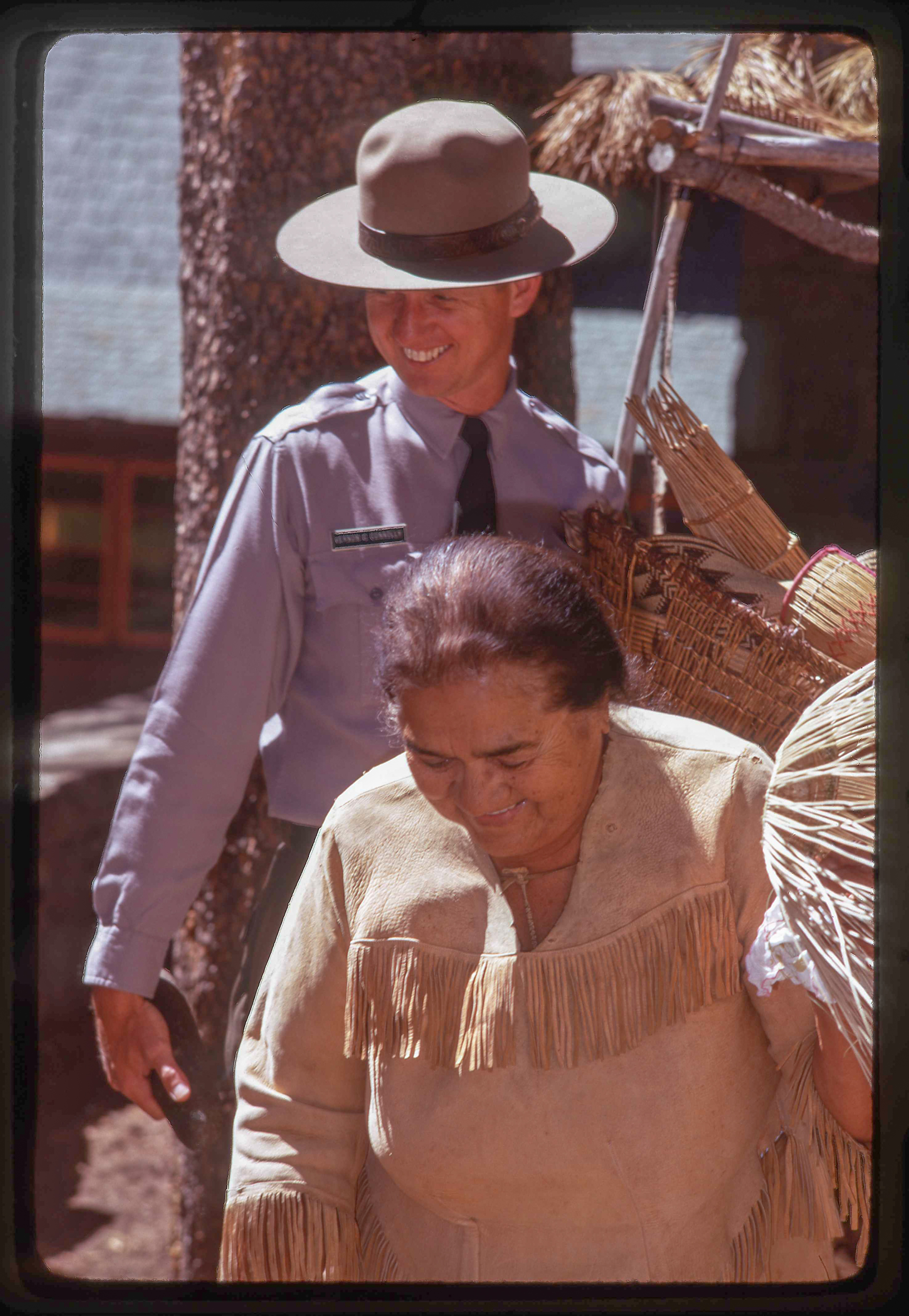 A woman in American Indian attire looks down,  a park ranger stands behind her holding baskets