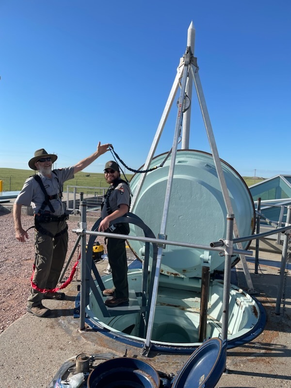 Two men in gray shirts and green pants standing in a gravel lot by an open hatch. The hatch is painting a seafoam green, surrounded by railings. 