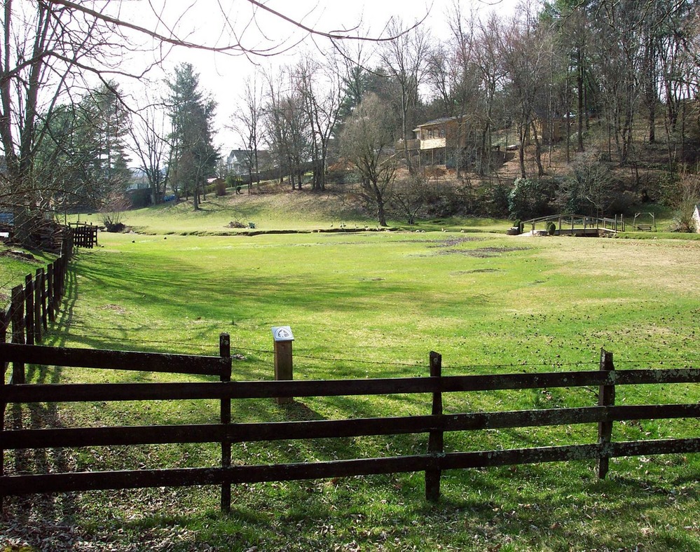 A green field inside a wooden fence with trees and homes visible in the background.
