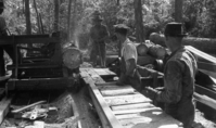 Historic black and white photo of 3 men working at a sawmill, watching as a log is sawed with a metal blade and sending sawdust in the air.