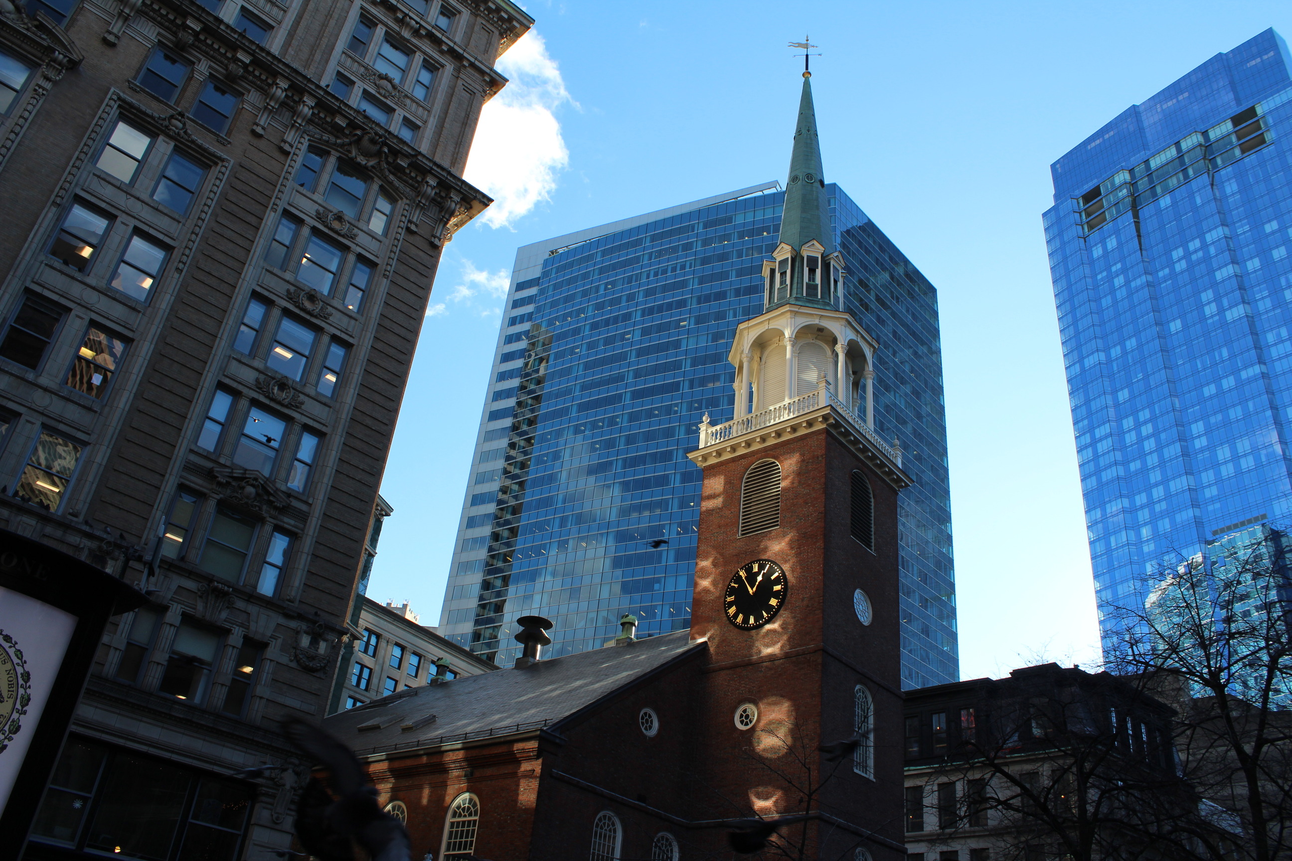 An image of Old South Meeting House taken from ground level and looking up. The building is made up of dark red brick and is shaped like a rectangle with a tower inserted at a short end. The tower has multiple windows and a clock on the side facing the photographer. There is a wooden balcony level on top of the brick and above that is a greening copper spire reaching into the blue sky. Surrounding the building are others of different ages and materials.