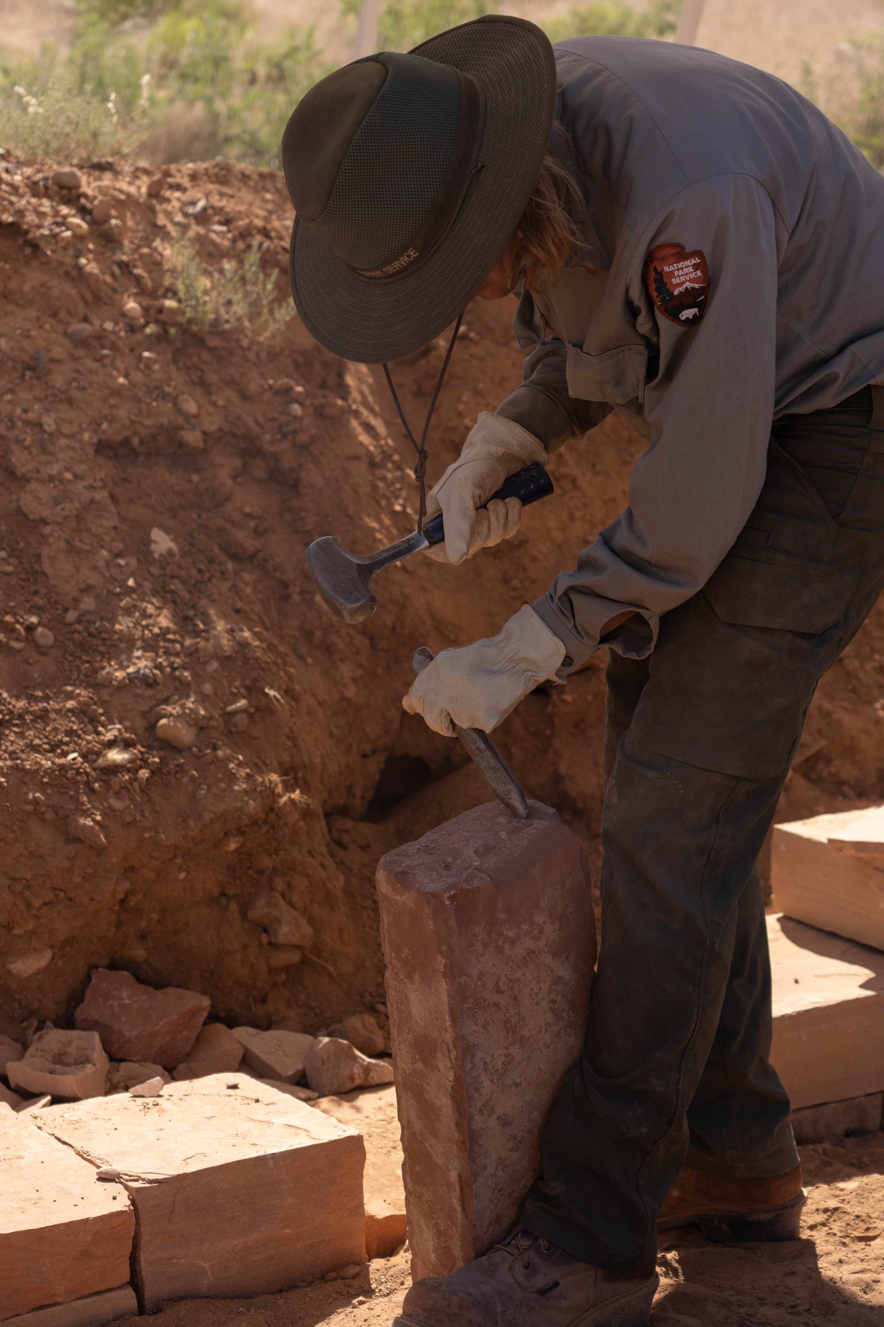 A person in green and grey working attire chiseling an orange sandstone brick near a dirt mound.