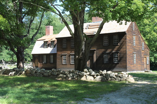  A large wooden colonial home in the saltbox design. Field stone wall in front of house along with several large shade trees. 