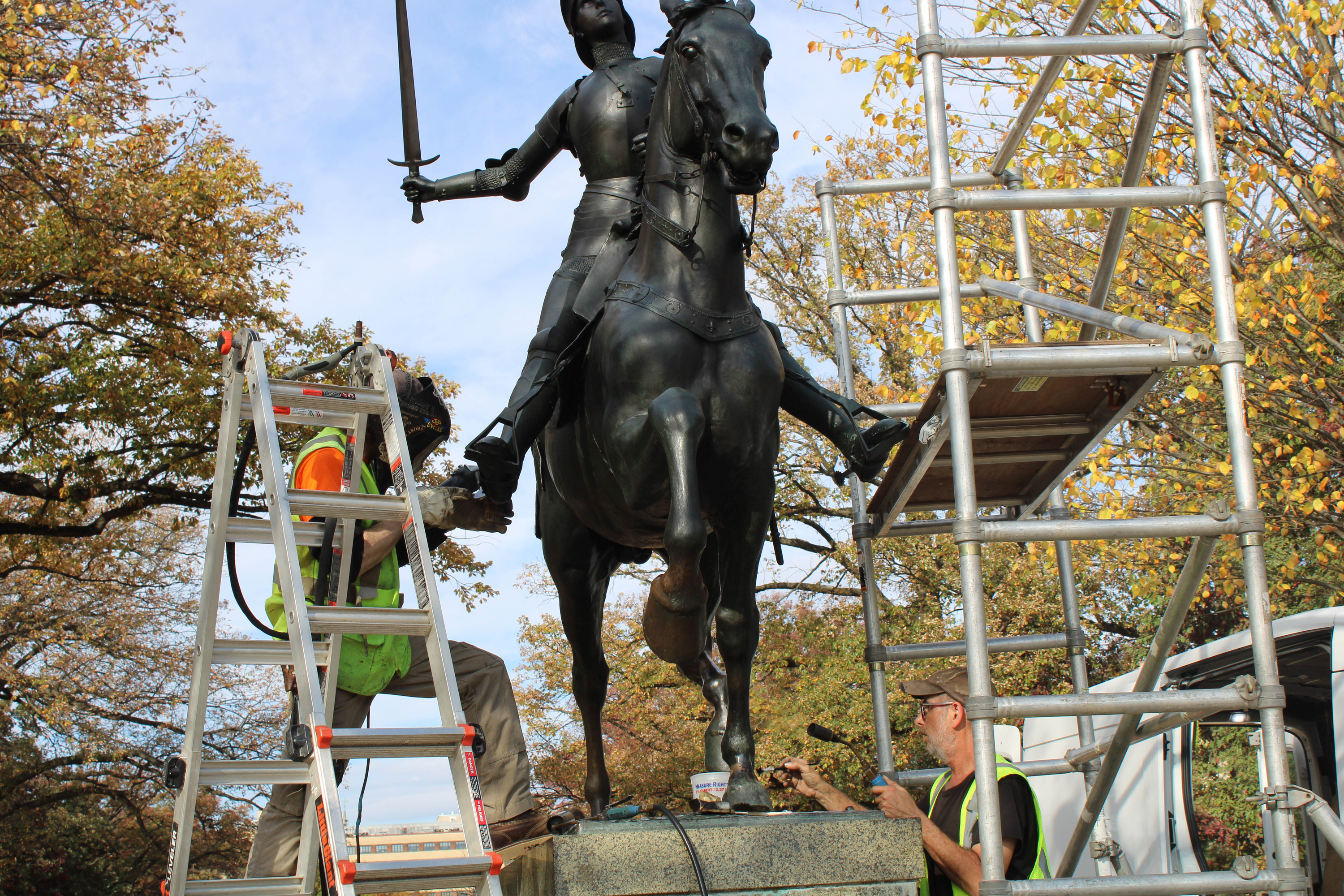 Two men stand on a ladder and scaffolding platform to perform maintenance on the Joan of Arc statue.