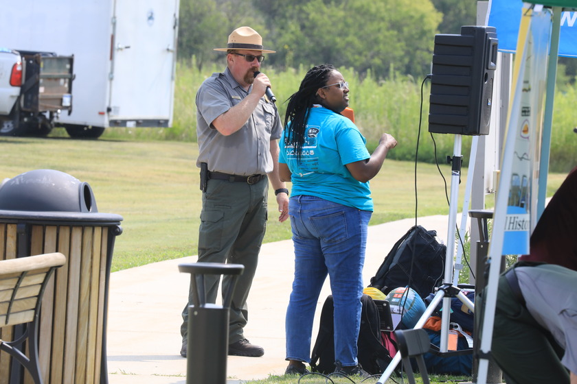 Uniformed NPS park ranger speaks into a microphone. A lady with a binder and shirt designating her role with the solar car challenge stands next to him. There is a sound system and tent nearby.