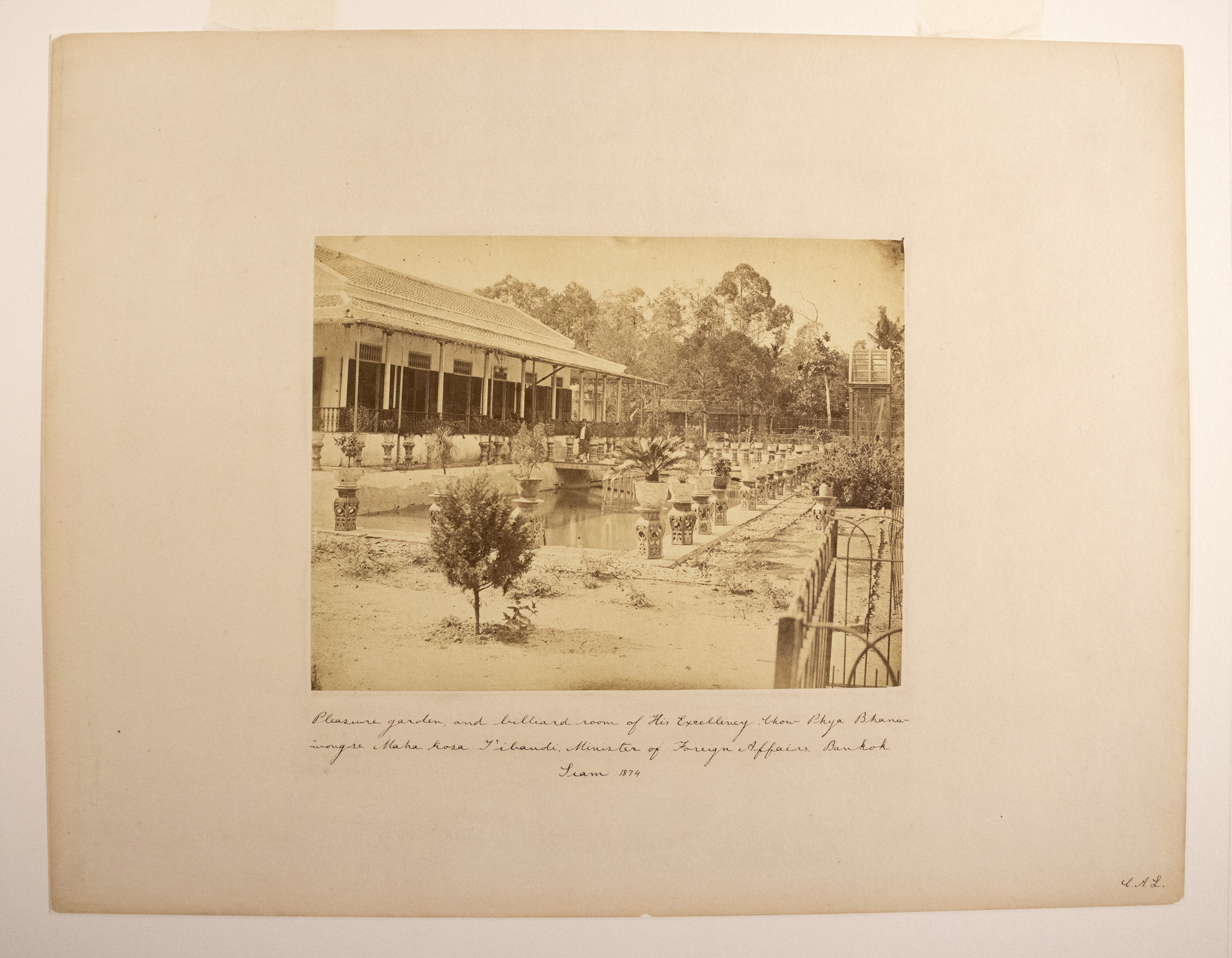 Sepia photograph with an inscription representing a garden with potted plants, a manmade pool with a bridge and a man standing on it, a building and trees in the background.