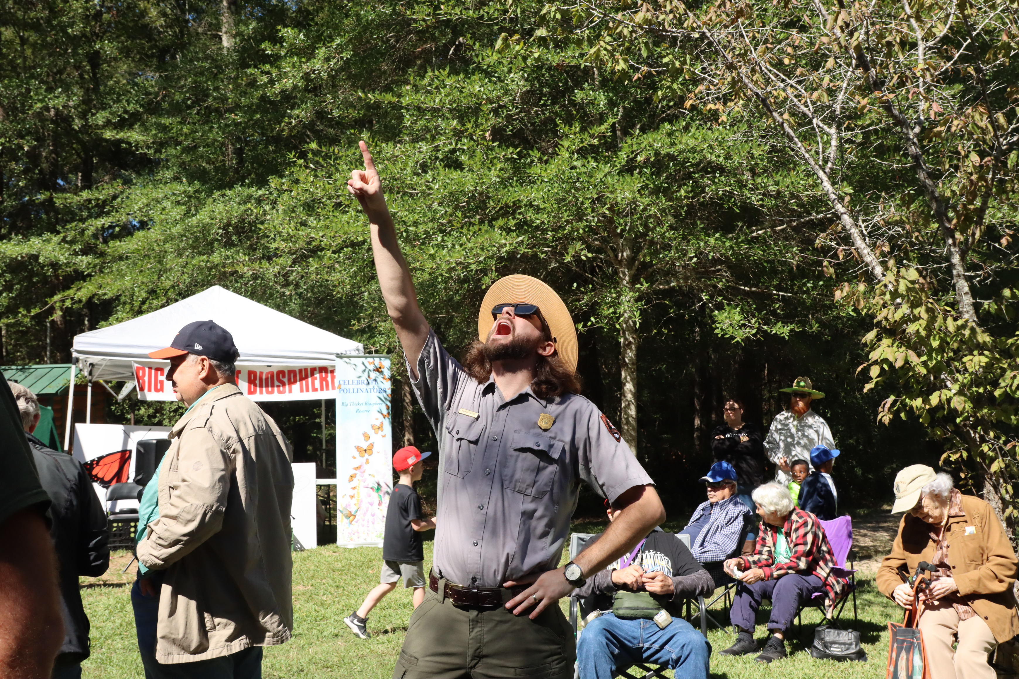 a park ranger wearing eclipse glasses with an open-mouth expression points upward while other people sit in chairs on the lawn in the background.