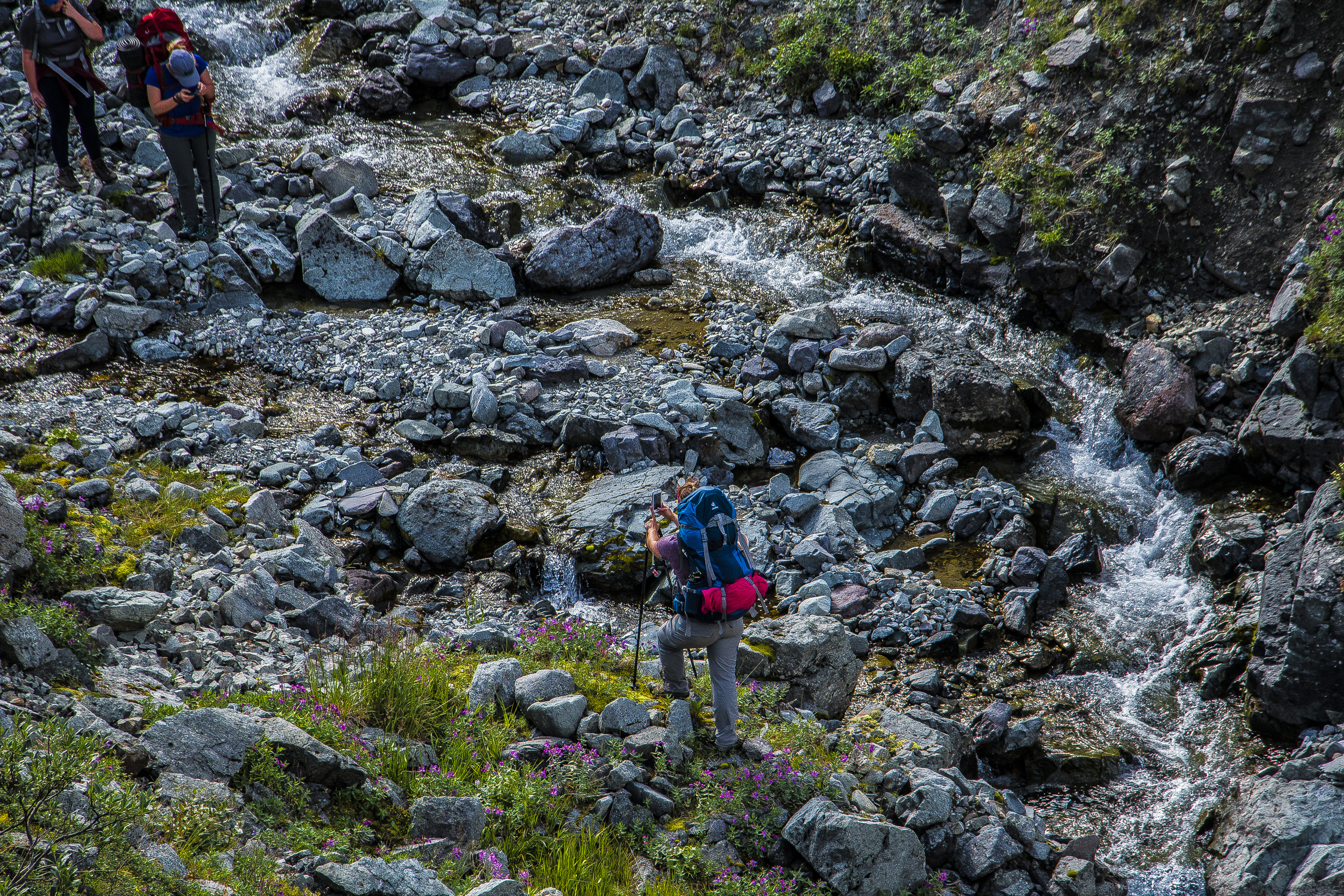 three people hiking up a rocky mountainside next to a shallow, swift creek
