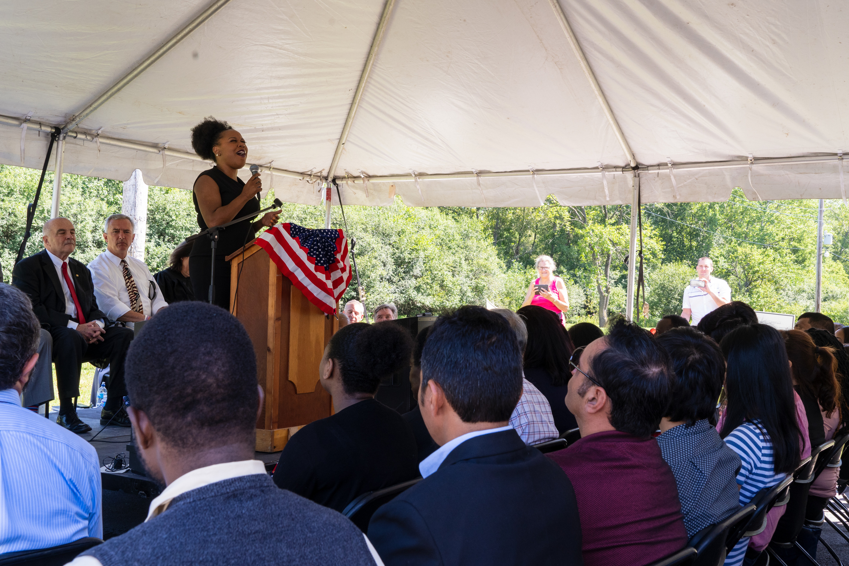 People listen to a woman singing at a podium under a tent.