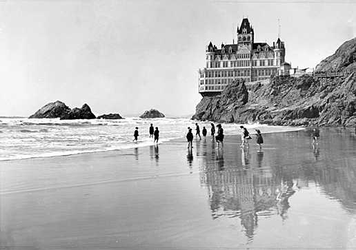 A dozen people wade ankle-deep in shallow water on a beach. Rocky outcrops and a large hotel on a cliff are behind them.