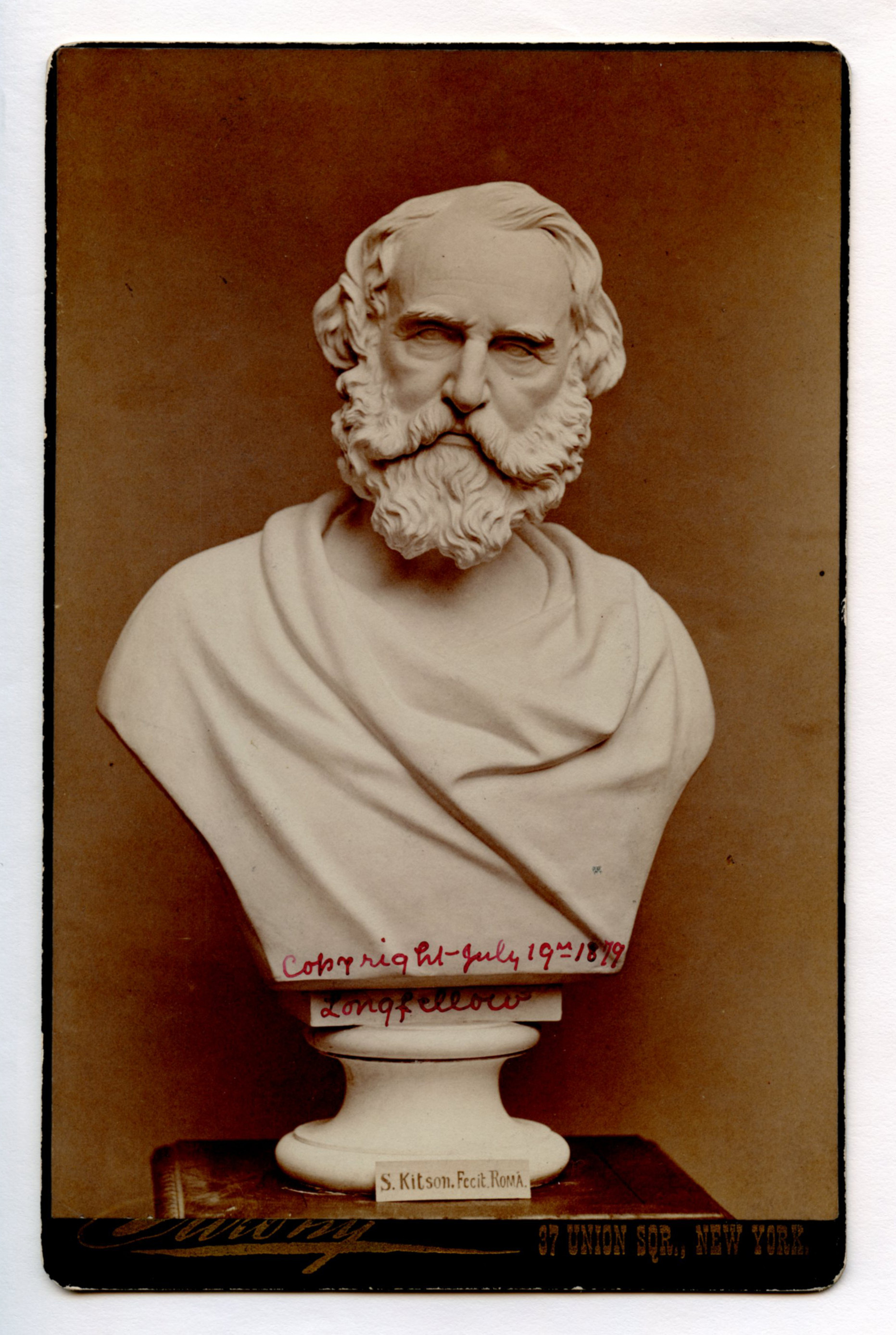 Photograph of Henry Wadsworth Longfellow portrait bust carved in white material. Bust is standing on a square table with label "S. Kitson. Fecit Roma" at the bust's base. Face, hair, and full beard are carved in detail. Chest and shoulders are carved to look like they're draped in cloth.
