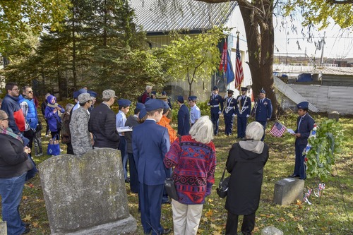 People standing in a cemetery, dressed warmly on a chilly autumn day, with flags and weraths visible.   