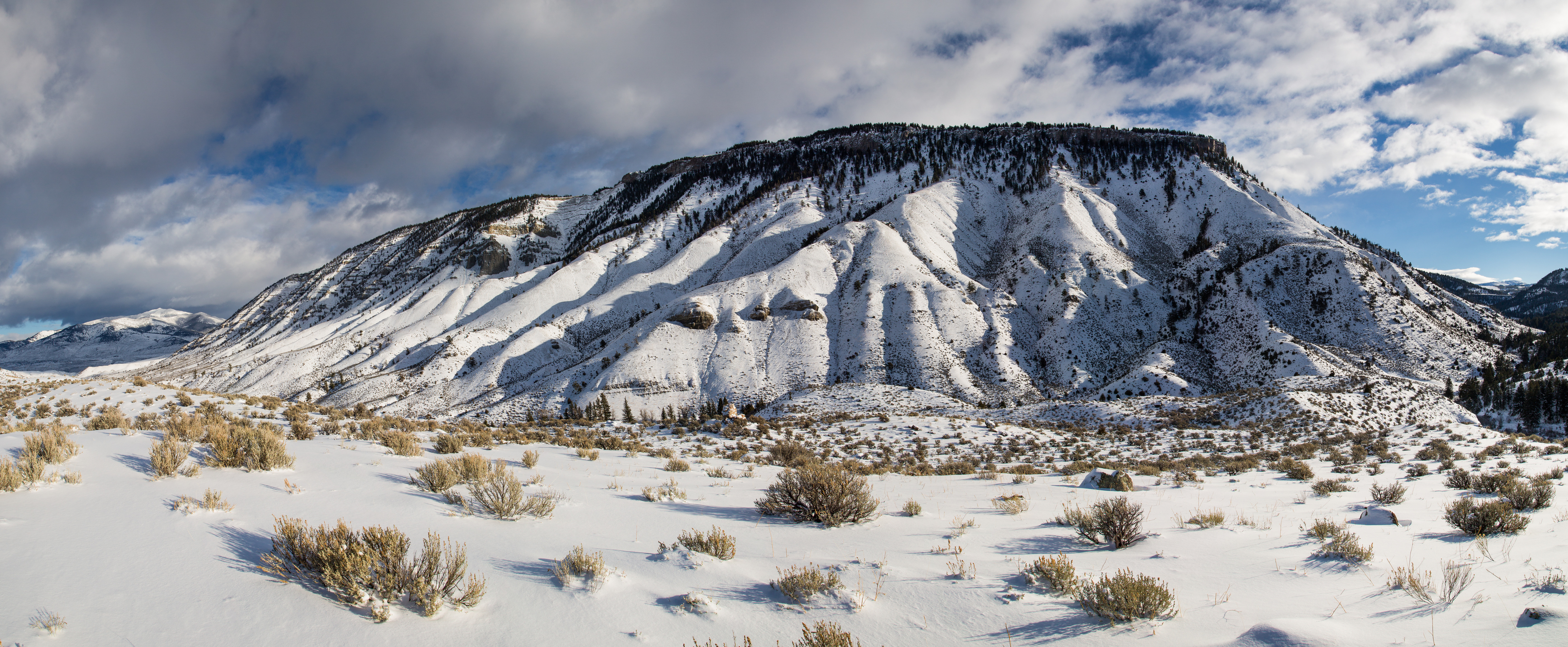 Looking across rugged, sagebrush, snow covered, flat at a snow covered, flat topped mountain.