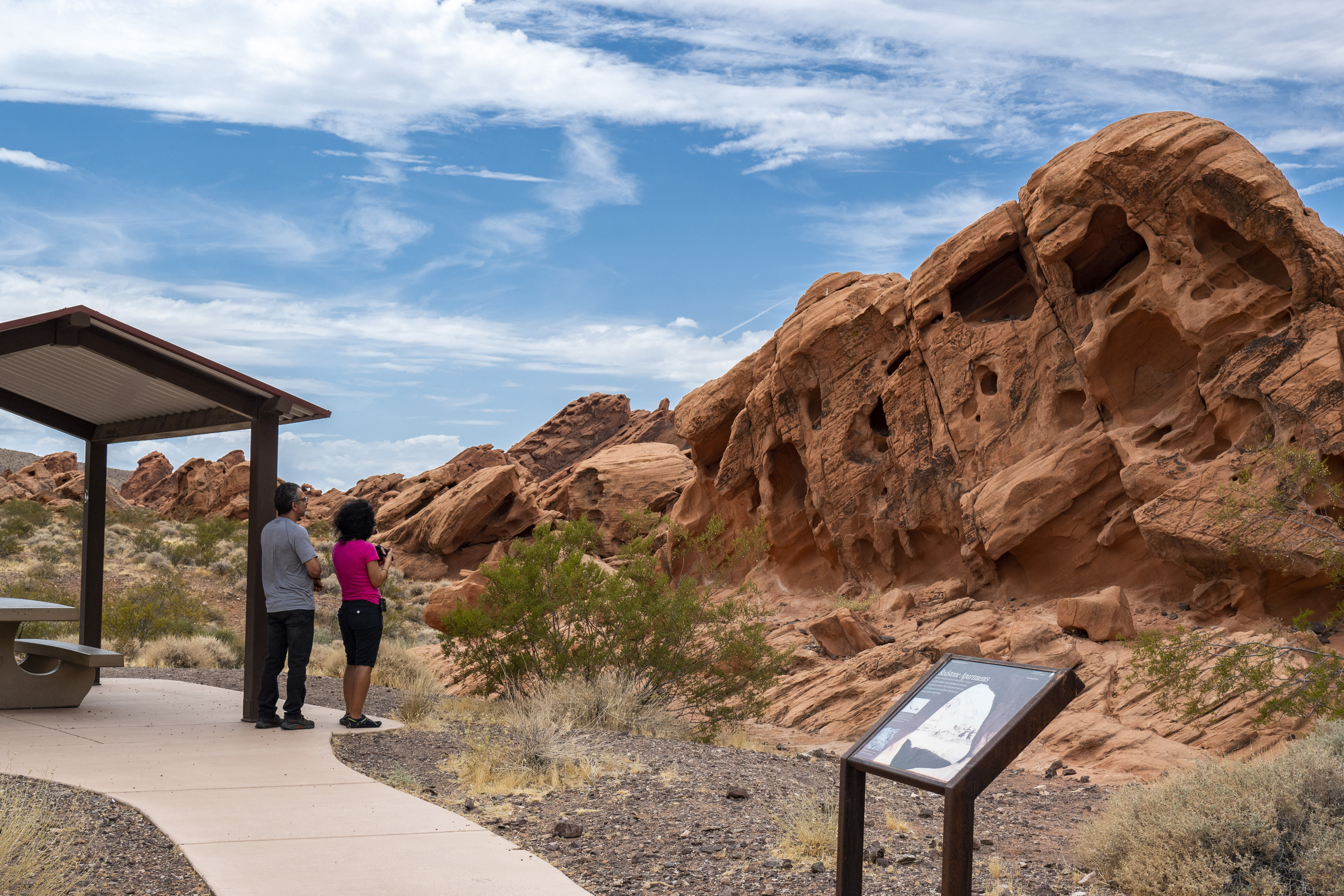 hikers at lower left stand near picnic shelter and educational wayside sign, large sandstone geologic formation to center and right, cloudy sky behind