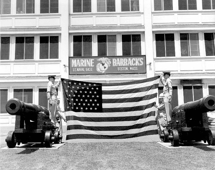 Two marines hold up a large American flag in front of the Marine Barracks. Two cannons are on either side of them. 