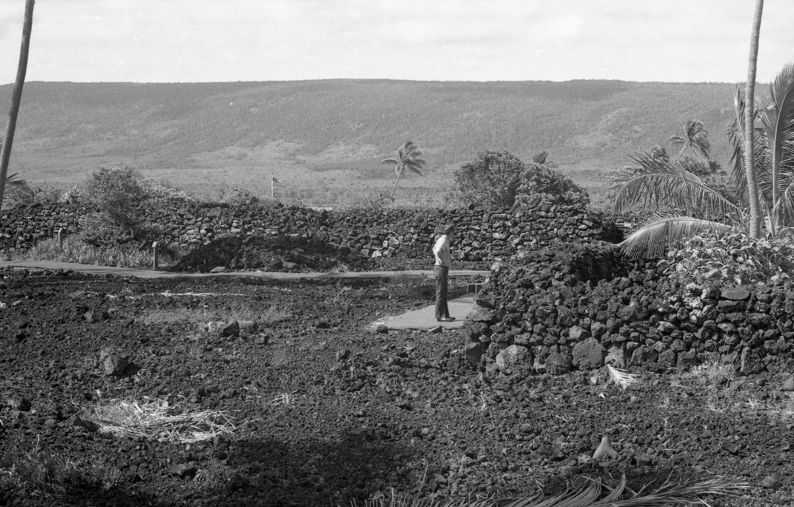 A black and white image of a National Park Service ranger. He is located towards the center right side of the image and is standing on a sidewalk. His body is turned towards the right side of the image and has his hands on his hips. His head is turned facing away from the camera. He is wearing a National Park Service uniform which includes a gray short sleeve shirt with two front pockets, green pants, and dress shoes. He is also wearing a ball cap with the national park service arrowhead logo embroidered onto the front of it. The lava rock wall starts on the left side of the image behind the park ranger then makes a turn to the right coming towards the camera. It then makes a left before trailing off the right side of the image. The sidewalk runs along the side of the wall and stops where the man is standing. On the left side of the image behind the sidewalk is a wooden post with a sign attached to it. Behind the lava rock wall is a flagpole with an American flag attached to it.