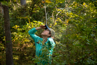 A biologist wearing a turquoise shirt holds her hat and a measuring tap as she looks up at the trees