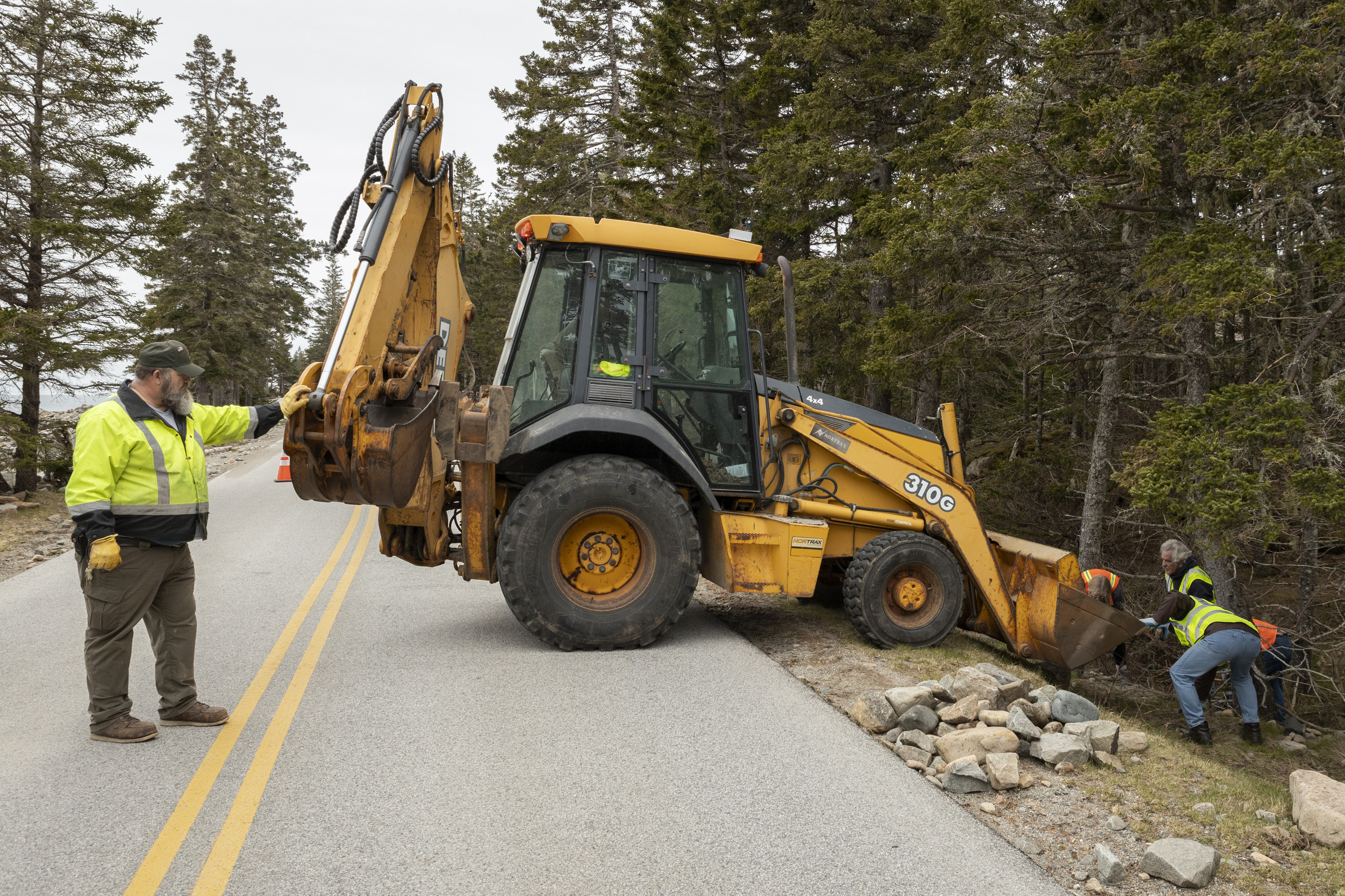 A backhoe and volunteers in high-visibility jackets working on a roadside rock cleanup in a forested area of Acadia National Park.