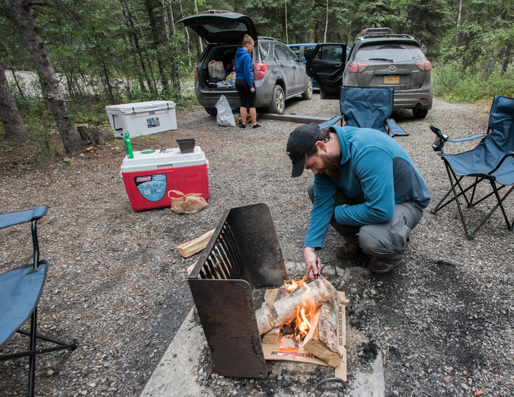 man building a fire in a grate near a woman removing items from the back of a car